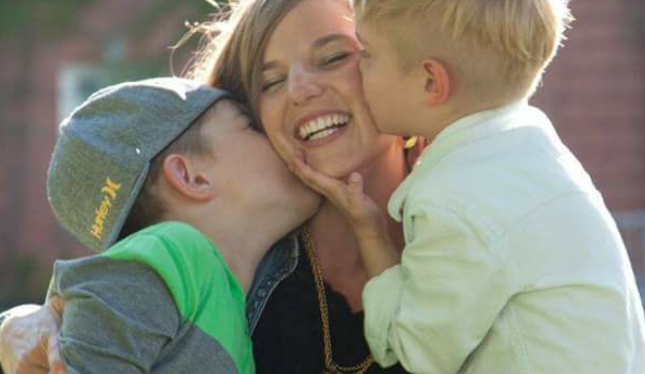 A photo of a mother smiling while two little kids kiss her on both cheeks.