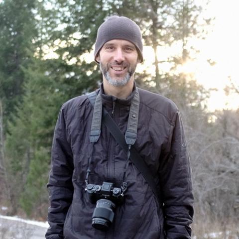 A middle-aged white man with a grey beard is smiling for the camera, wearing a grey beanie, and has a professional camera around his neck 