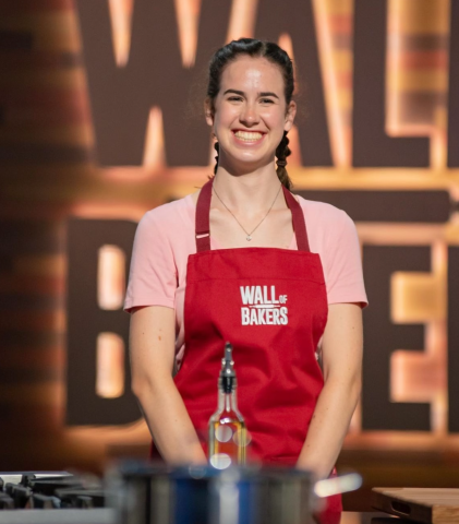 Photo of Mackenzie Tomlin, on the "Wall of Bakers" Food Network TV show. She is a young white woman with dark brown hair in two braids, wearing a pink t-shirt and a red apron that reads "Wall of Bakers". 