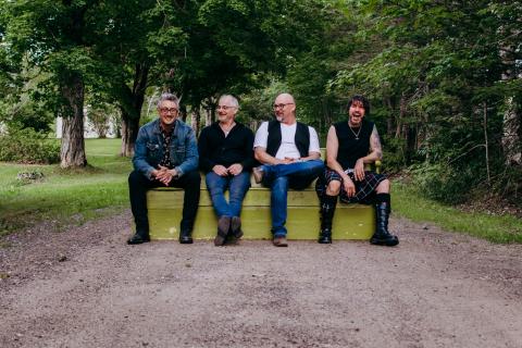A photo of Joker's Right band. Four people are sitting on a bench on a dirt road outside. They are surrounded by green trees