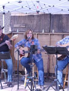 "Two in the Morning Trio": Two older white men and a middle-aged white woman sit under a tent playing guitar and singing, all wearing country-esque attire