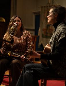 "The Dusty Halos": Two mid-late twenties white women sit by a microphone and sing and play guitar