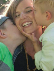 A photo of a mother smiling while two little kids kiss her on both cheeks.