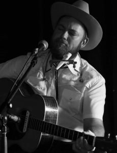 Black and white image of a middle-aged white man in his mid 30s, Zakary Miller, performing on a stage and playing the guitar