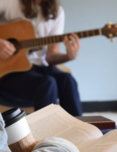 A photo of a man reading a book with a to go coffee cup. There are also two men playing guitar behind him. 